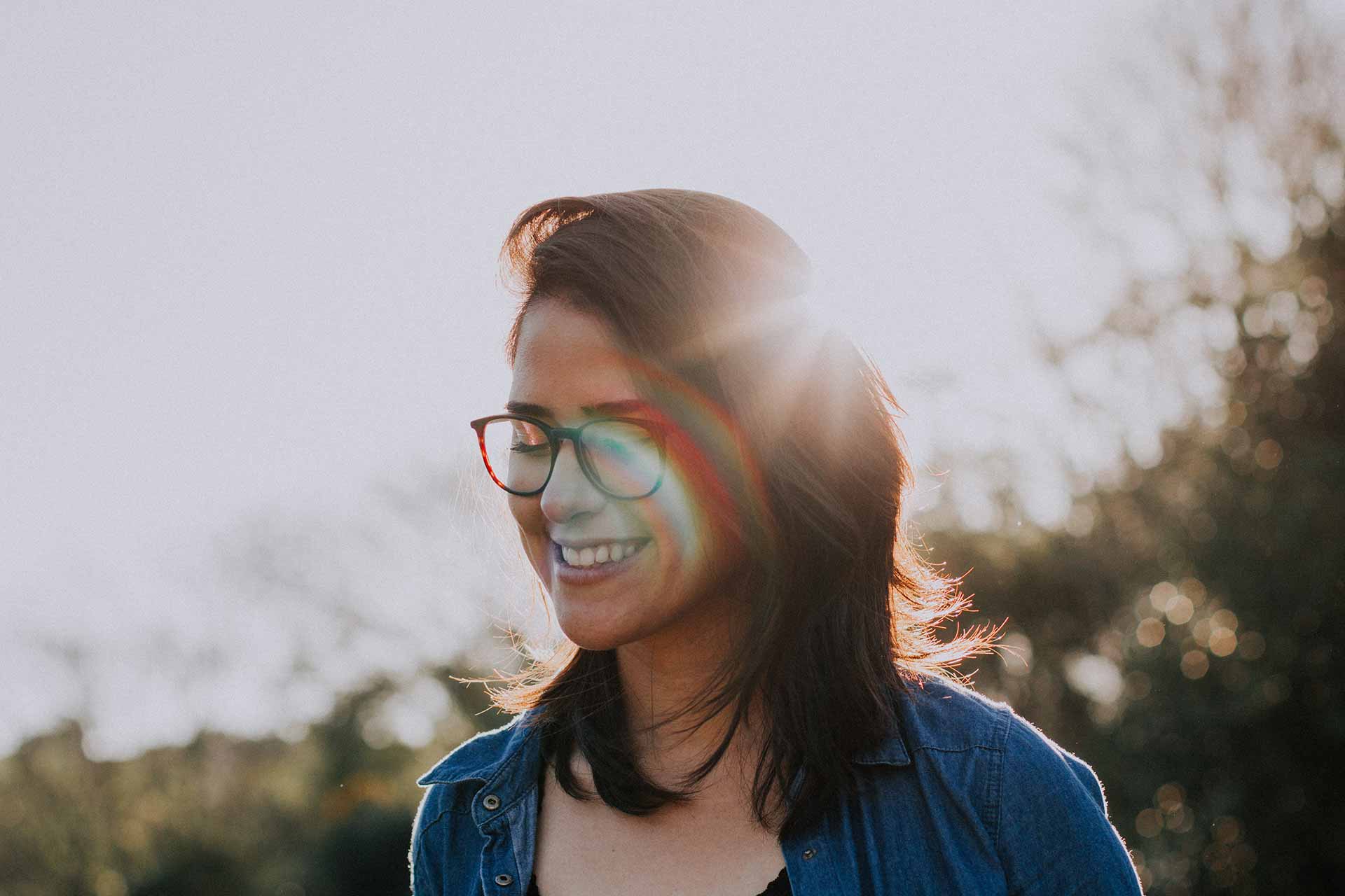 Smiling woman with glasses outdoors bathed in warm, soft sunlight.